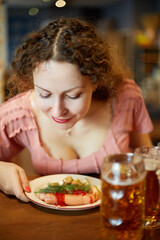 Smiling woman with curly hair sniffs fried sausages with sprig of dill on plate in cafe
