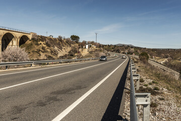 A two-lane road with one lane in each direction with a solid white line in the middle with some vehicles driving and a metal guardrail