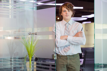 Handsome teenager in shirt and tie poses near glass door in modern office