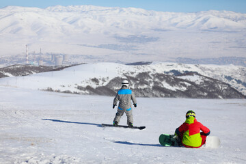 Two snowboarders are on mountain in ski resort, back view, factory far away