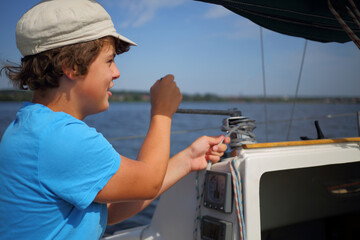 Boy teenager in cap controls boat during sailing on river at summer day