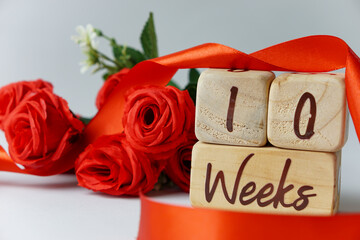 10 week gestational age milestone written on a wooden cube with red roses and ribbons, and a white...