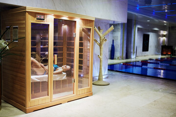  Woman in swimsuit lies on bench in infrared sauna cabin behind glass doors in recreational area in...