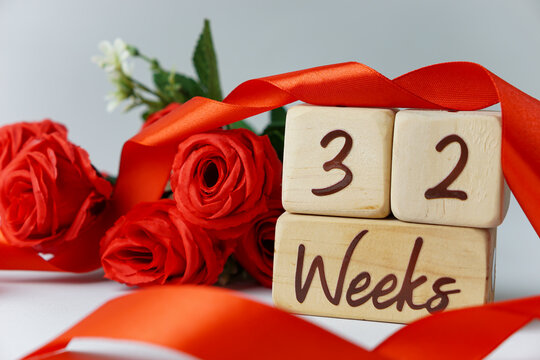 32 week gestational age milestone written on a wooden cube with red roses and ribbons, and a white background