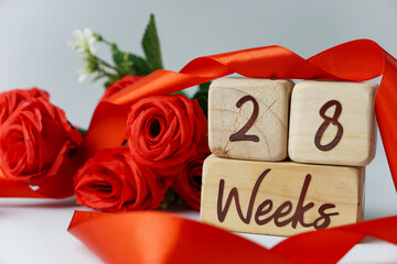 28 week gestational age milestone written on a wooden cube with red roses and ribbons, and a white background