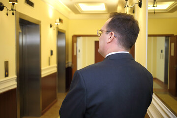 Fototapeta premium Man in a business suit standing in the hallway between the elevator and mirrors in the luxury apartment building, back view