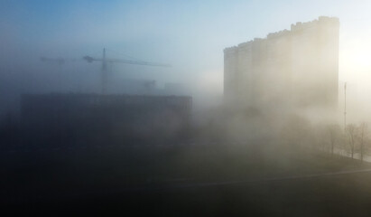 Construction cranes and a multi-storey residential building, shrouded in fog, in the early spring morning.