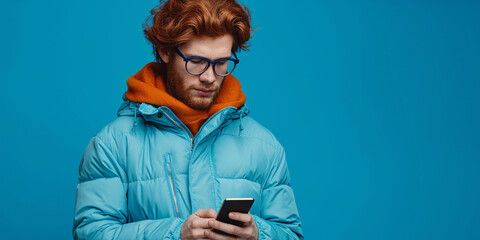 Stylish man with red hair wearing glasses and blue winter jacket using smartphone on blue background - Powered by Adobe