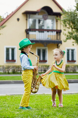 Little boy in dancing suit plays saxophone and little girl dances on grassy lawn against two-storied house