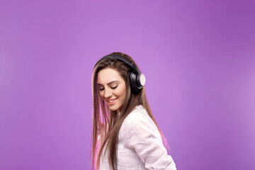 Beautiful young woman in wireless headphones listening to music and dancing on  purple background. Girl uses wireless earphones