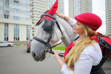 Coachman woman stands with horse in red harness near residential buildings
