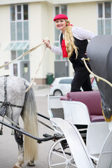Happy coachman blonde on trestles holds reins on couch with horse near residential buildings