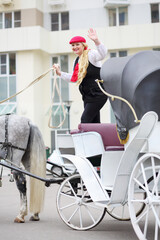 Happy coachman blonde on trestles waves hands on couch with horse near residential buildings