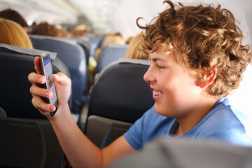 Handsome teenager boy with curly hair holds ohone in airplane during travel