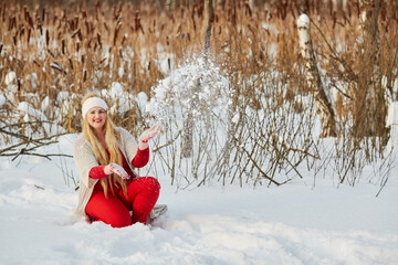 Smiling woman sits squatted in snowbank and throws snow up in winter park