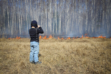 The little boy with the action camera on his head looking at a burning dry grass along the birch...