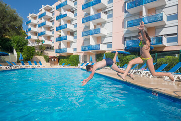 Happy boy and girl jump in pool near hotel at summer sunny day - obrazy, fototapety, plakaty