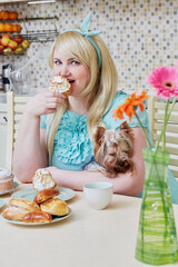 Plump smiling woman holding little dog sits eating homemade baked sweets at table in kitchen