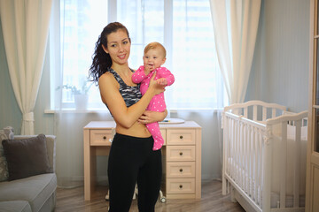 Mother with curly hair in sportswear holding a baby in front of a window in the room - obrazy, fototapety, plakaty