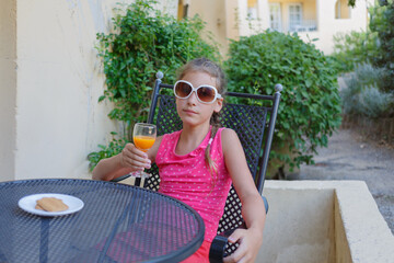 Girl in pink t-shirt and sunglasses with orange juice and biscuit sitting at table