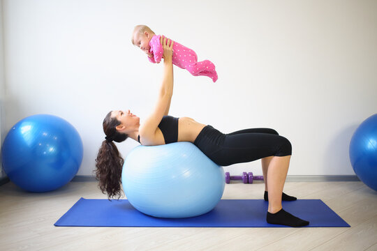 Happy woman lying on the big blue ball on her back lifted up smiling baby in the gym