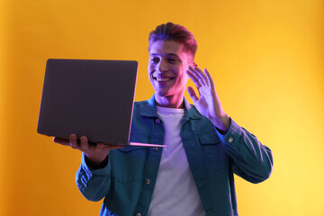 Young man with laptop talking via video chat on yellow background