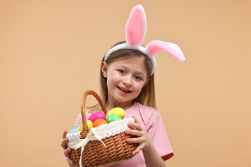 Easter celebration. Cute girl with bunny ears holding basket of painted eggs on beige background