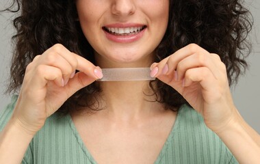 Young woman holding teeth whitening strip on grey background, closeup