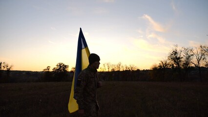 Young male military in uniform walks with flag of Ukraine at meadow. Soldier of ukrainian army going with blue-yellow banner on field at sunset. Concept of victory against russian aggression. Slow mo - 755758737