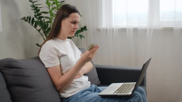 Wellness and dieting concept. Upset young caucasian woman using computer sitting alone on sofa at home, holding pills tablet pack read prescription medicine label about vitamin information online