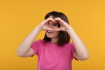 Happy woman looking through folded in shape of heart hands on orange background