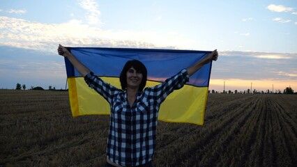 Happy woman looks into camera standing on wheat field with a lifted blue-yellow banner at sunset. Ukrainian smiling lady posing with a raised over head flag of Ukraine on barley meadow at sunrise. - 755760361
