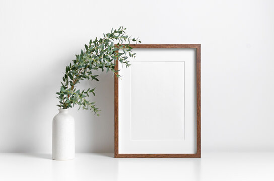 Wooden vertical picture frame mockup in white interior with fresh eucalyptus plant in vase
