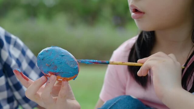 Happy children in park, cute asian girl and boy with mix race friend, close up hand paint egg with paintbrush together on green grass in garden. Kids celebrate Easter holiday outdoor