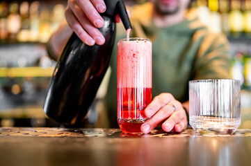 A bartender pours a vibrant red cocktail into a glass at a modern bar, with focus on the drink and hands. Bottles of alcohol line the background, creating an intimate atmosphere.