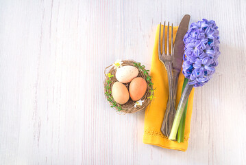 Festive place setting for easter holiday dinner. Vintage fork and knife, blue hyacinth flower on yellow napkin and birdnest with eggs on white wooden background Space for text, top view