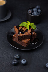 Chocolate brownie cake with mint, blueberries, chocolate topping and espresso. Traditional American dessert. Popular cake with dark chocolate on a black background. Close-up, selective focus.