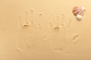 Fototapeta na wymiar Honeymoon concept. Handprints, seashells and two golden rings on sand, top view
