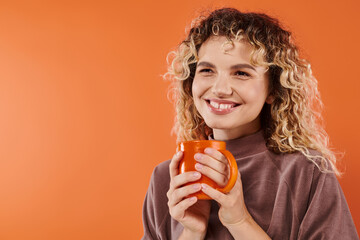 cheerful woman with curly hair and cup of morning coffee looking away on orange, daily routine