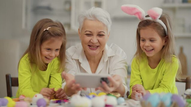 Easter grandmothers with granddaughters. Smiling grandmother with twins grandchildren watching cartoons on phone in rabbit bunny ears, talking, preparing together at home. Happy Easter concept.