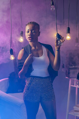 Fashionable ethnic woman. Girl stands among light bulbs hanging on wires. African American woman looking at camera. Cute girl in smoke. African American woman in trendy clothes. Female millennial