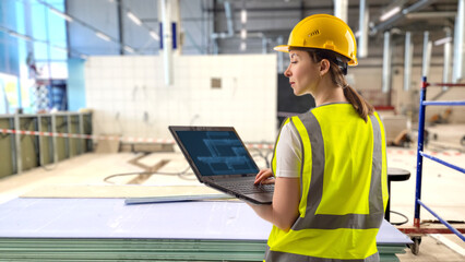 Woman builder with laptop. Engineer stands in building under construction. Girl inspector monitors progress of construction. Woman builder in yellow hardhat and vest. Lady builder designer