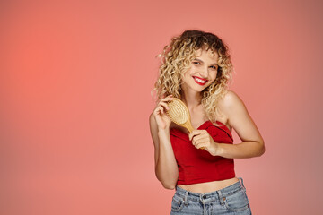 charming and curly woman in red trendy top posing with hair brush on yellow and pink backdrop