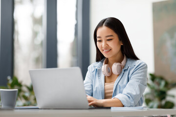 Smiling asian freelancer woman with headphones working on laptop
