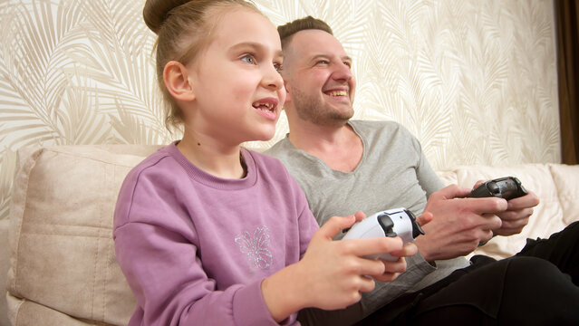 A father spends time with his little daughter, sitting on the sofa and playing a game console. Weekend activities, free time, home entertainment and video games concept.