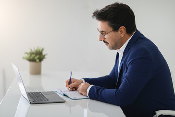 Dedicated businessman in a sleek blue suit taking notes in a planner beside his laptop