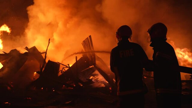 Firefighters bravely spray water on a massive fire in a rural market, using high-pressure hoses to extinguish the intense flames. Safety and disaster response are critical as they battle the blaze