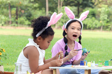 Cute Asian girl with curly hair African friend wearing bunny ears, painting egg with paintbrush together on green grass meadow in garden. Kids celebrating Easter holiday outdoor Happy children in park
