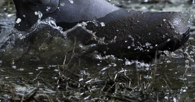 Super slow motion macro of farmer in rubber boots walks through mud puddles with splashing water drops after rainy day on green agricultural natural food production farmland fields at 1000 fps.