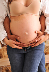 Close up photo of pregnant woman in white shirt and blue jeans and her husband holding their hands on pregnant belly. The concept of happy parenthood